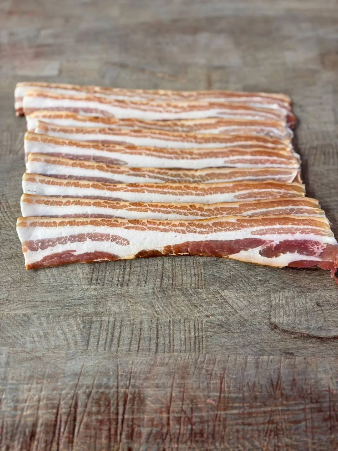 Rashers of black streaky bacon laid in a row on a worn wooden chopping block