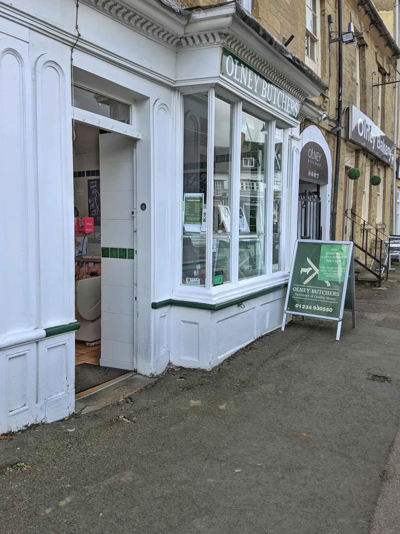 The bay window of Olney Butchers from the pavement, with the green plinth line along the base and the green A-board showing the shop's logo